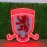 A red and white illuminated LED neon sign of the Middlesbrough Football Club crest, displayed on a wooden surface against a green ivy wall background.