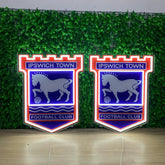 Two illuminated Ipswich Town Football Club neon-style wall signs displayed side by side in front of a green artificial foliage backdrop.