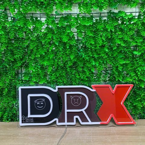 A 3D acrylic LED sign spelling out 'DRX' with a black, dark brown, and red color scheme, set against a green artificial ivy wall background.