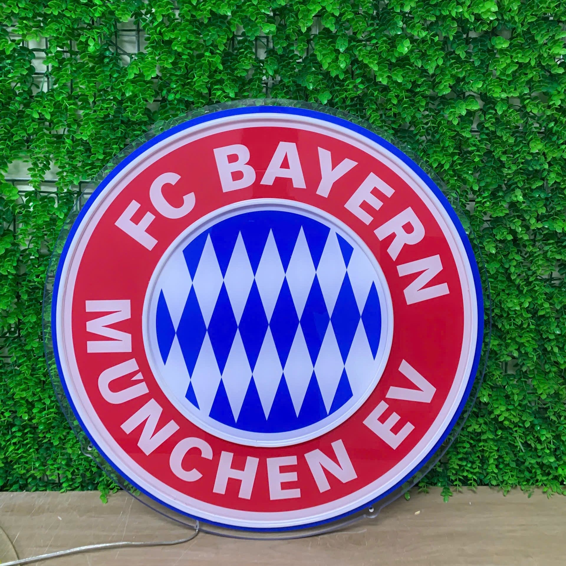 A large circular FC Bayern München EV sign featuring the club's red, white, and blue logo, displayed against a green ivy-covered wall.