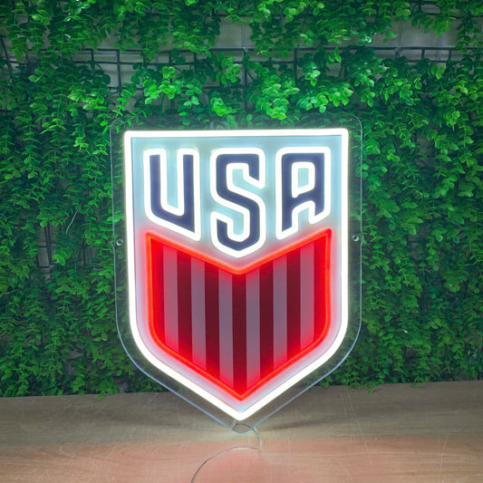 A shield-shaped USA soccer logo sign with a glowing white border, navy blue 'USA' lettering, and red and white vertical stripes, displayed against a green leaf wall.