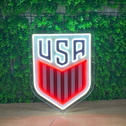 A shield-shaped USA soccer logo sign with a glowing white border, navy blue 'USA' lettering, and red and white vertical stripes, displayed against a green leaf wall.