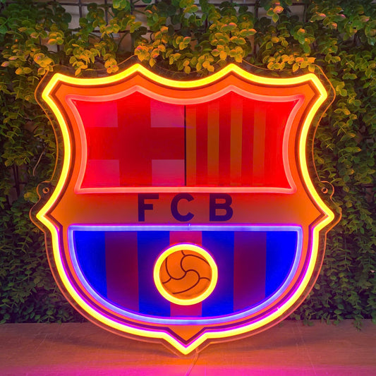 A large FC Barcelona neon sign glowing brightly against a background of green ivy-covered walls. The illuminated crest features red and blue stripes, yellow accents, and the letters 'FCB' in the center.