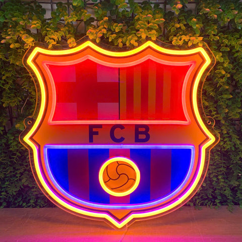 A large FC Barcelona neon sign glowing brightly against a background of green ivy-covered walls. The illuminated crest features red and blue stripes, yellow accents, and the letters 'FCB' in the center.